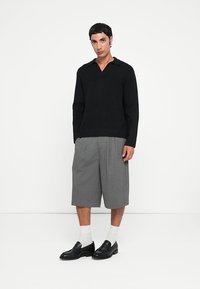 Maglione nero a maglia con scollo a V e orlo a costine, abbinato a pantaloncini grigi a pieghe e mocassini neri sopra calze bianche.