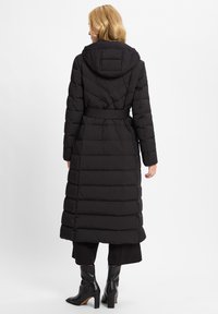 Vrouw met blond haar die een lange zwarte pufferjas met capuchon, zwarte broek en zwarte haklaarzen draagt, staande tegen een witte achtergrond.