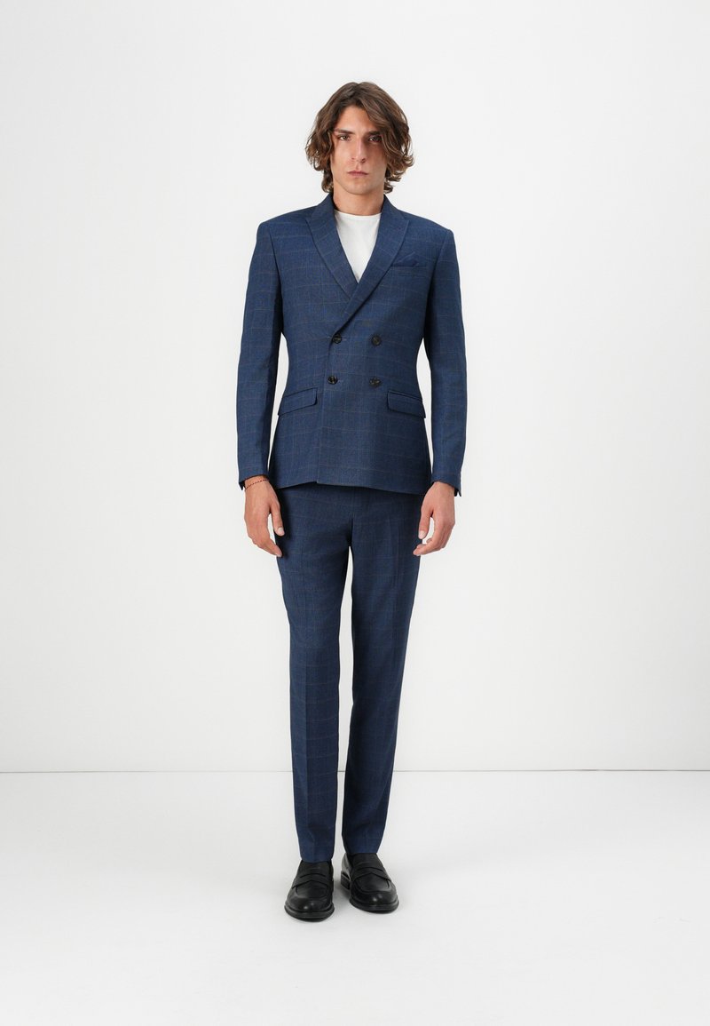 Costume bleu marine à double boutonnage avec motif écossais, en tissu texturé. Design ajusté, associé à une chemise blanche et des chaussures noires.