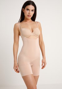 Beige shapewear bodysuit met een gladde, rekbare textuur, voorzien van verstelbare schouderbanden en een lengte tot halverwege de dij. Uitsparing bij de borst.
