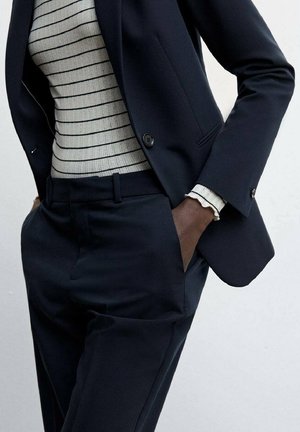 Personne portant un costume bleu marine, les mains dans les poches, et un pull blanc côtelé avec des rayures horizontales noires.