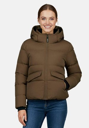 Femme souriante portant une veste matelassée marron avec fermeture éclair à l’avant et poches à rabat, associée à un jean bleu, debout avec une main dans la poche.