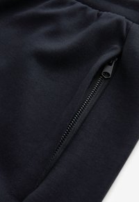 Jogging pantalon noir en tissu lisse, présentant une poche à zip inclinée sur le côté pour un rangement sécurisé.