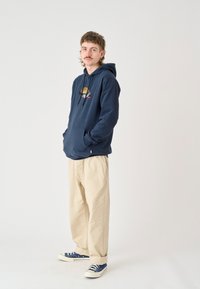Navy hoodie with kangaroo pocket and embroidered graphics, paired with light beige loose-fitting trousers and blue sneakers.