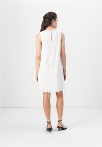 Robe blanche sans manches avec un motif texturé, un ourlet festonné et un détail en forme de trou de clé au dos. Portée avec des talons hauts noirs à lanières.