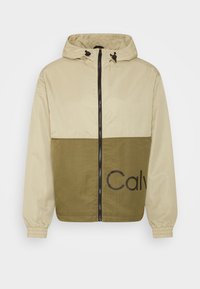 Beżowo-oliwkowa kurtka w stylu color block z zamkiem b�łyskawicznym, regulowanym kapturem, elastycznymi mankietami i wyraźnym logo „Calv” na dolnej części.