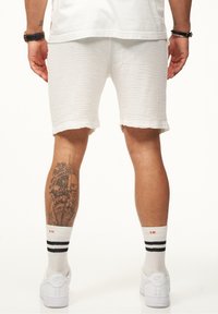 Weiße strukturierte Shorts mit fransigem Saum, kombiniert mit weißen Sneakers und gestreiften weißen Socken. Tattoo am Unterschenkel sichtbar.
