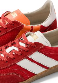 Paire de baskets basses rouges et blanches avec des accents orange, des lacets rouges, un tissu texturé et des rayures blanches sur les côtés.