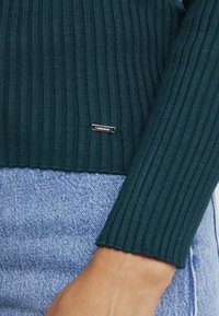 Geribbelde, donkergroene trui met een hoge halslijn en gedetailleerde manchetten. Klein zilveren label geplaatst bij de zoom. Gedragen over lichtblauwe denim.