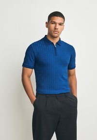Blauw poloshirt met een patroonstructuur, korte mouwen en een ritssluiting bij de kraag, gecombineerd met een zwarte broek.