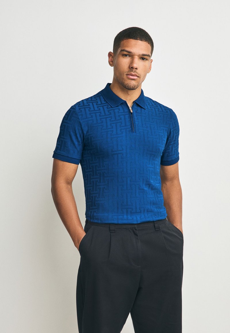 Blauw poloshirt met een patroonstructuur, korte mouwen en een ritssluiting bij de kraag, gecombineerd met een zwarte broek.