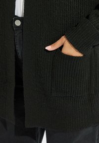 Svart stickad cardigan med en texturerad, ribbad mönster, med framfickor och långa ärmar. Bärs över mörka byxor.