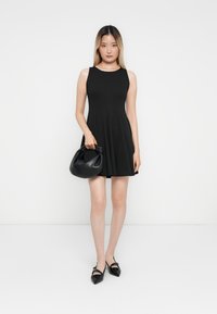 Robe noire sans manches avec une jupe évasée, associée à un petit sac à main en cuir noir et des ballerines noires brillantes à bout pointu. Fond blanc uni.