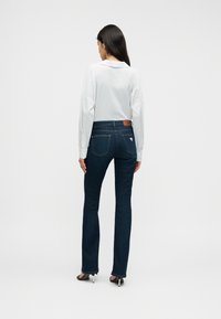 Σκούρο μπλε καμπάνα τζιν με υφή από τζιν ύφασμα, με καφέ δερμάτινο έμβλημα στη μέση και διακριτικές πίσω τσέπες.