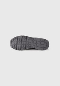 Semelle de chaussure en caoutchouc noir avec un motif de relief texturé, comportant des rainures et des sections surélevées pour une meilleure adhérence. Marque estampillée sur le côté.