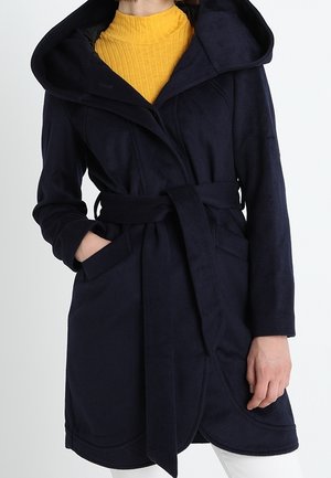 Woman in a navy blue belted coat with hood and long sleeves, wearing a mustard yellow ribbed turtleneck underneath.
