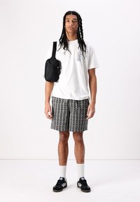 T-shirt blanc avec logo, short noir à motifs, sac bandoulière noir, chaussettes blanches, baskets noires. Modèle se tenant contre un fond uni.