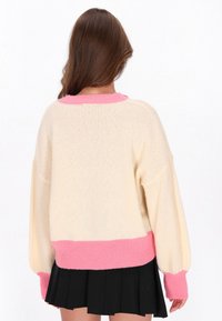 Donna con lunghi capelli castani che indossa un maglione crema con polsini e orlo rosa, abbinato a una gonna plissettata nera, di spalle.