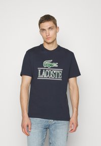 Camiseta de algodón azul marino con mangas cortas, featuring un gran logo verde de Lacoste centrado en el pecho y franjas horizontales blancas debajo del texto.