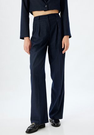 Pantalons bleu marine à rayures avec taille haute et plis, en tissu lisse ; associés à des mocassins noirs brillants.