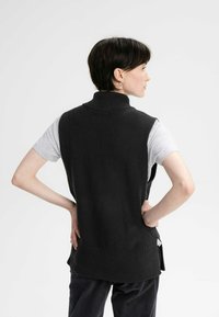 Schwarze Strickweste mit hohem Kragen, getragen über einem grauen T-Shirt. Die Weste hat eine lockere Passform mit Seitentaschen und einem strukturierten Muster.