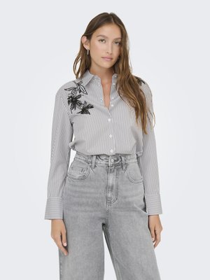 Stripet skjorte med knapper som har svart blomsterbroderi på skulderen; parret med høyt liv grå denim jeans.