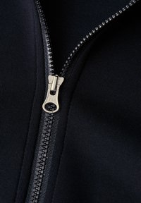 Tela negra con una textura suave y cuenta con un zipper metálico YKK. El primer plano muestra los dientes del zipper y el detalle de la lengüeta.