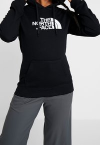 Felpa nera con cappuccio e logo bianco "THE NORTH FACE" sul davanti. Presenta una tasca a marsupio e cappuccio con cordini. Tessuto morbido, vestibilità casual.