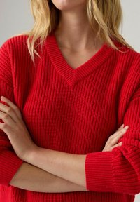 Czerwony sweter dzianinowy z dekoltem w serek, z wyraźnym prążkowanym wzorem i szerokimi mankietami. Materiał wydaje się gruby i przytulny.