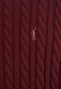 Bordeaux kabelgebreide trui met dikke gedraaide patronen en een klein beige geborduurd figuur op de voorkant.