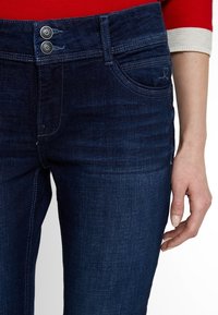 Jeans in denim blu scuro con chiusura a due bottoni, cinque tasche e una leggera texture. Il tessuto appare resistente e liscio.