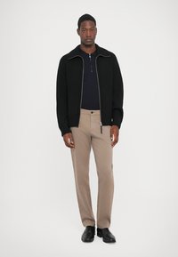 Mann iført svart zip-up genser, marineblå skjorte, beige bukser og svarte dressko, stående mot en lys ensfarget bakgrunn.