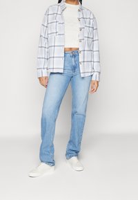 Kvinna i ljusblå jeans, vit texturerad crop top, rutigt skjort överdel och vita sneakers som står mot en neutral bakgrund.