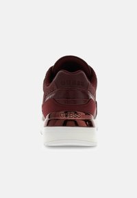 Basket maroon avec des matériaux lisses et texturés, présentant un talon en cuivre glossy, une semelle blanche, et un logo "GUESS" en relief à l'arrière.