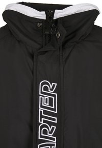Jacket negra aislante con capucha forrada en blanco, con cierre central y un gran logo bordado en blanco en el lateral.