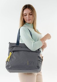 Donna con un maglione verde chiaro che porta una grande borsa tote grigia con trama, con manici blu navy e cursore della cerniera giallo, sulla spalla.