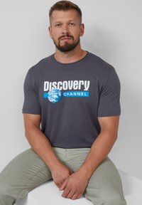 Grijze t-shirt met de tekst "Discovery CHANNEL" in het wit en blauw, met een wereldbolafbeelding. Korte mouwen en ronde hals. Draagt bij lichte groene broek.