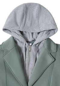 Veston vert superposé à un sweat à capuche gris clair zippé, capuche relevée avec cordons visibles.