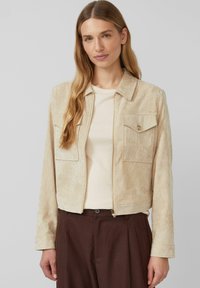 Beżowa kurtka z korduroju z kołnierzykiem, dwoma przednimi kieszeniami i zapięciem na zamek. Posiada długie rękawy i skrócony fason.