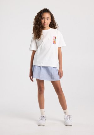 Jonge vrouw met krullend haar, gekleed in een wit t-shirt met opdruk, een lichtblauwe gestreepte rok, witte sokken en witte sneakers, tegen een effen achtergrond.
