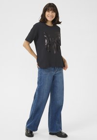 Schwarzes T-Shirt mit glänzendem "DANCE IT OUT"-Text, kombiniert mit lockeren blauen Jeans und schwarzen Lacklederschuhen. Flacher Hintergrund.