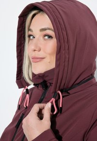 Žena upravující šňůrku kapuce vínové bundy, usmívající se a dívající se do strany.