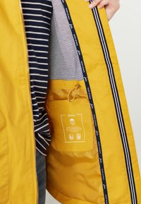 Wnętrze żółtej wodoodpornej kurtki z granatowo-białą paskowaną podszewką. Posiada kieszeń ze ściągaczem oraz nadrukowane etykiety z informacjami o marce.
