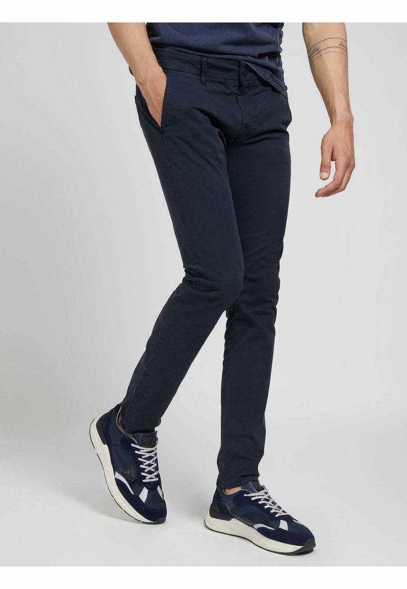 Pantaloni slim-fit blu navy in misto cotone con tasche frontali, zip e passanti per cintura, abbinati a sneakers atletiche blu navy.