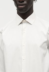 Κοντινό πλάνο ενός ατόμου που φοράει λευκό πουκάμισο με κουμπιά και μυτερό γιακά σε απλό φωτεινό φόντο.