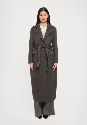 Woman wearing a long, dark brown belted coat over patterned wide-leg trousers and black pointed shoes, standing against a plain white background.