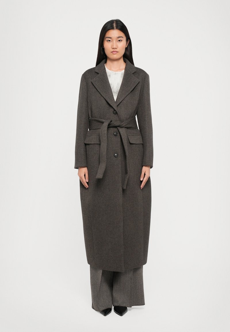 Femme portant un long manteau marron foncé ceinturé sur un pantalon large à motifs et des chaussures noires à talons pointus, se tenant devant un fond blanc uni.