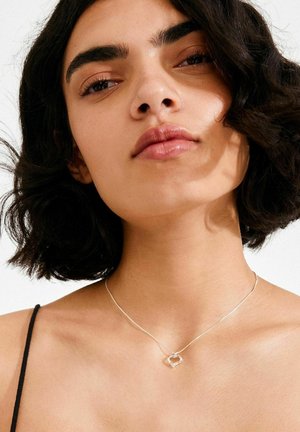 Ogrlica s obeskom v obliki srebrnega srca na tanki verigi, nošena na goli koži, z enostavno in moderno zasnovo na nevtralnem ozadju.