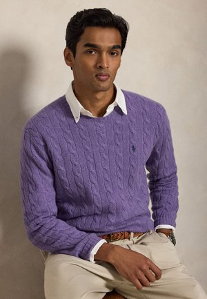 Jeune homme portant un pull en maille torsadée violet par-dessus une chemise blanche à col, un pantalon beige et une ceinture tressée marron, assis sur un fond uni.