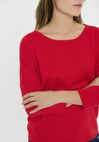 Červený, ryhovaný dlhý rukáv top s širokým okrúhlym výstrihom a voľným strihom. Rukávy majú na manžetách mierny rozširujúci sa tvar, čo poskytuje textúrovaný vzhľad.